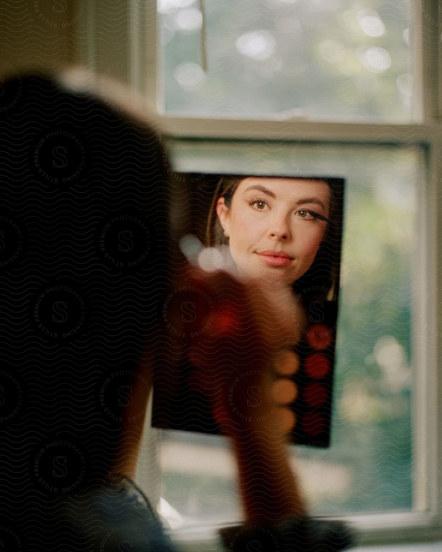 A woman with red lipstick applies mascara while gazing at her reflection in a mirror with eyeshadow cosmetics below.