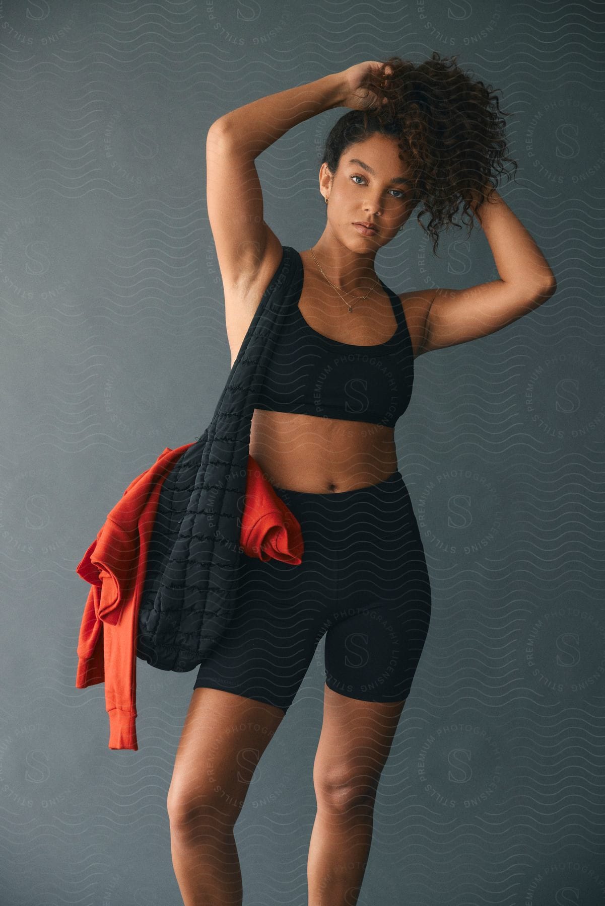 A young woman with curly hair poses while wearing fitness apparel and carrying a bag.