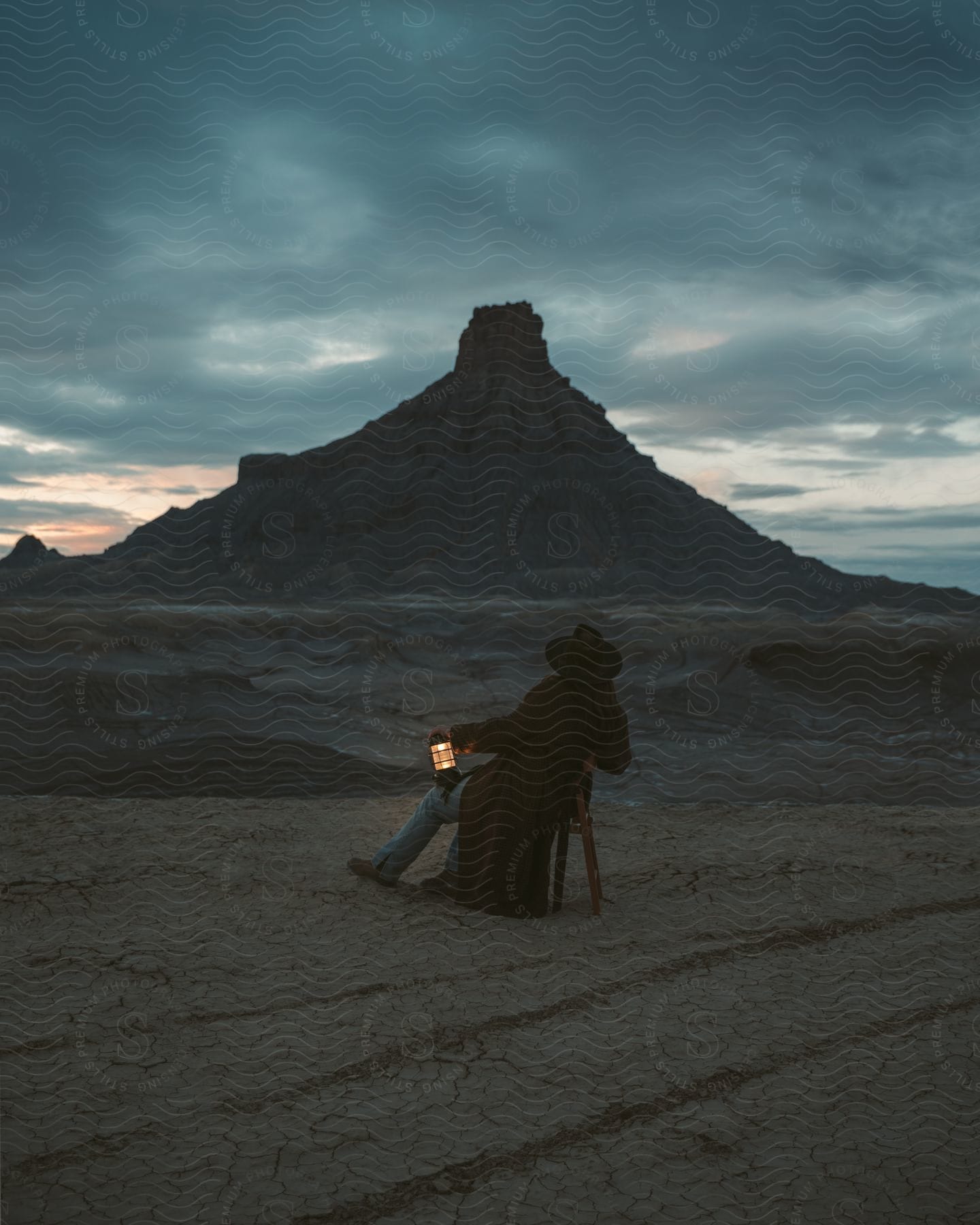 Man With A Black Cowboy Hat An Overcoat And Jeans Sits In Armchair In The Desert Watching Rock Formation In The Distance