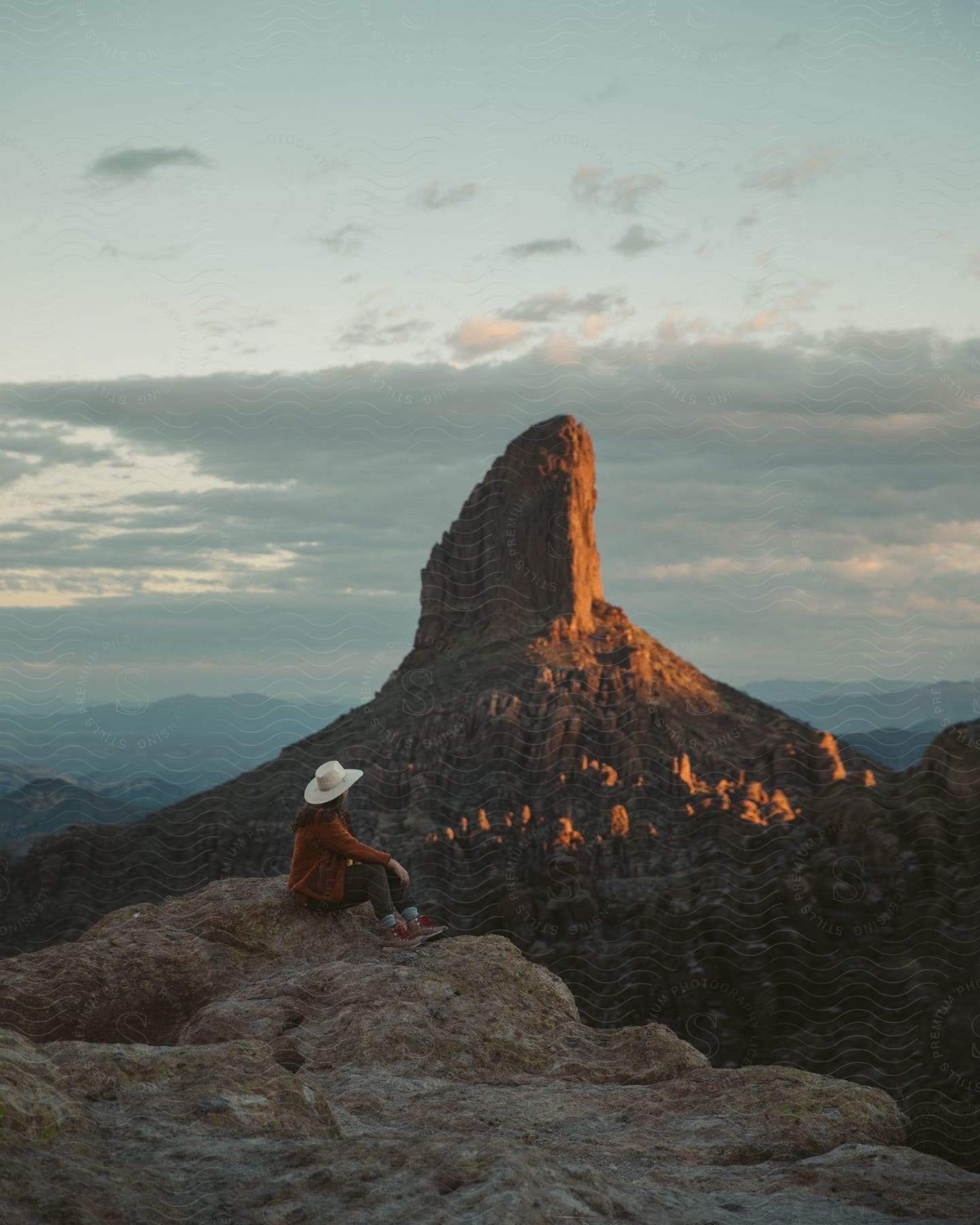 A woman wearing a cowboy hat sits on a ledge overlooking mountains and rock formations