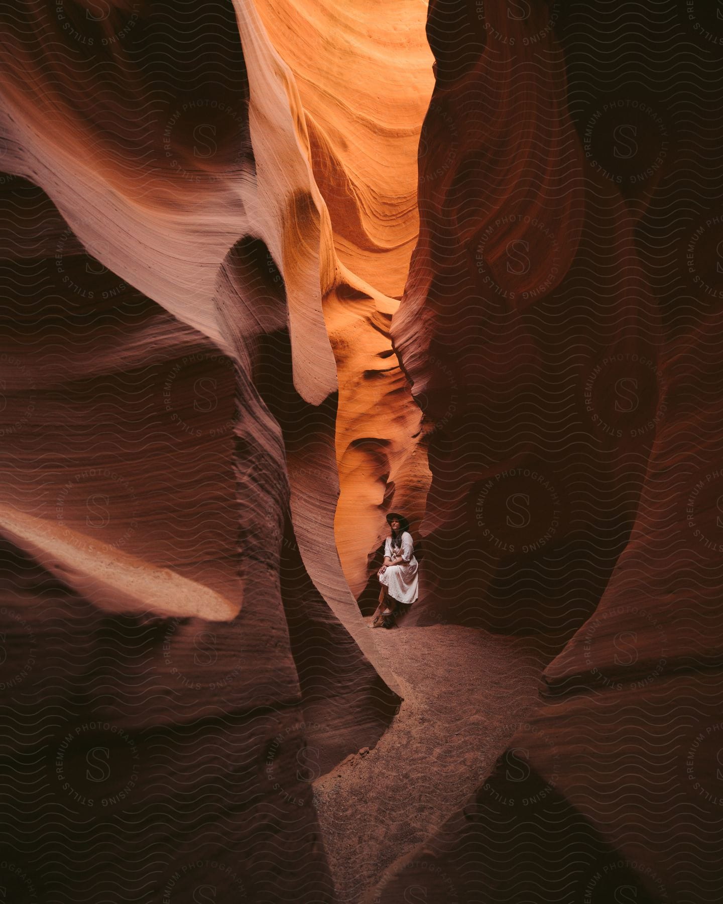 A woman wearing a hat and white dress is sitting in the middle of a slot canyon, with sunlight streaming in from above.