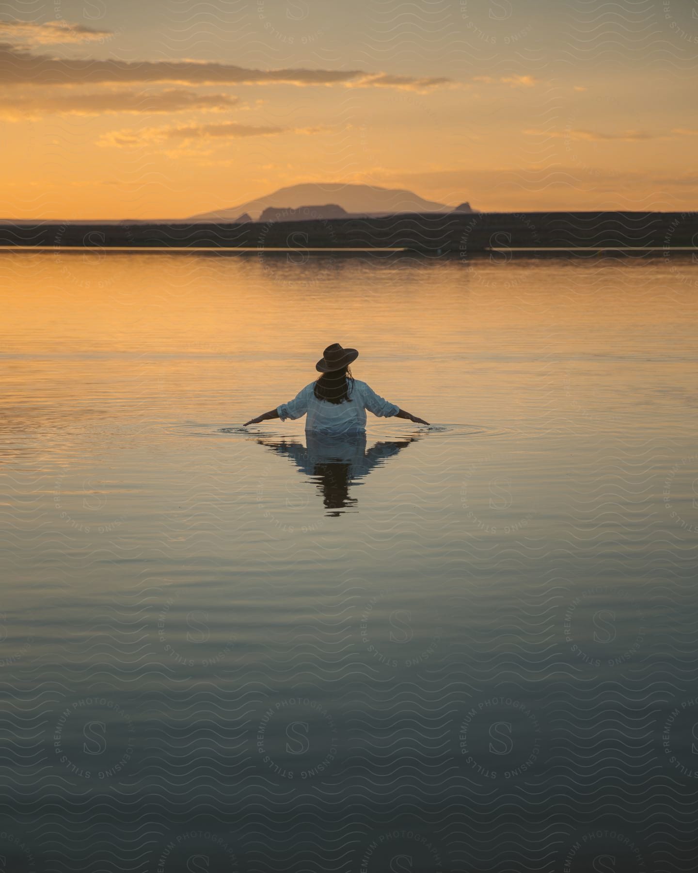 woman wearing a beige hat and a white shirt goes into the water at sunset