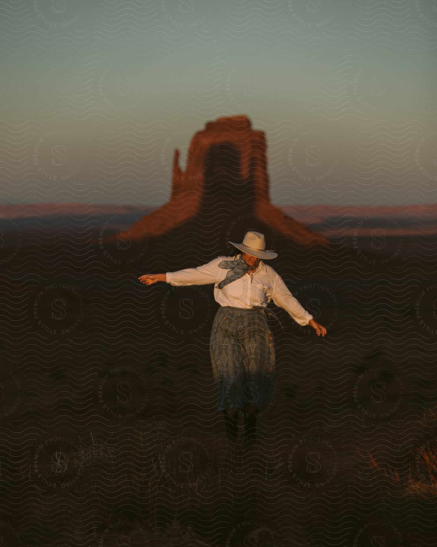 A cowgirl in a patterned dress skirt and cowboy boots stands before the West and East Mitten Buttes.