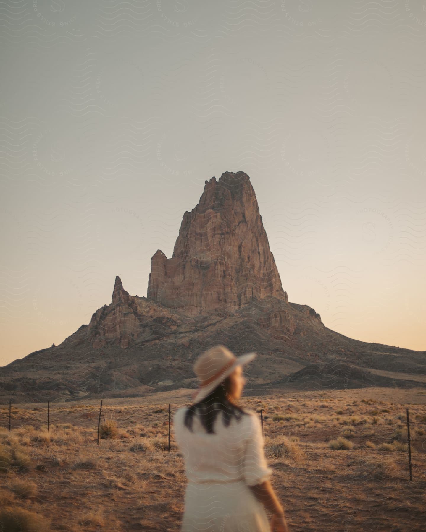 A woman wearing a cowboy hat is walking toward a fence with a mountainous rock formation in the distance