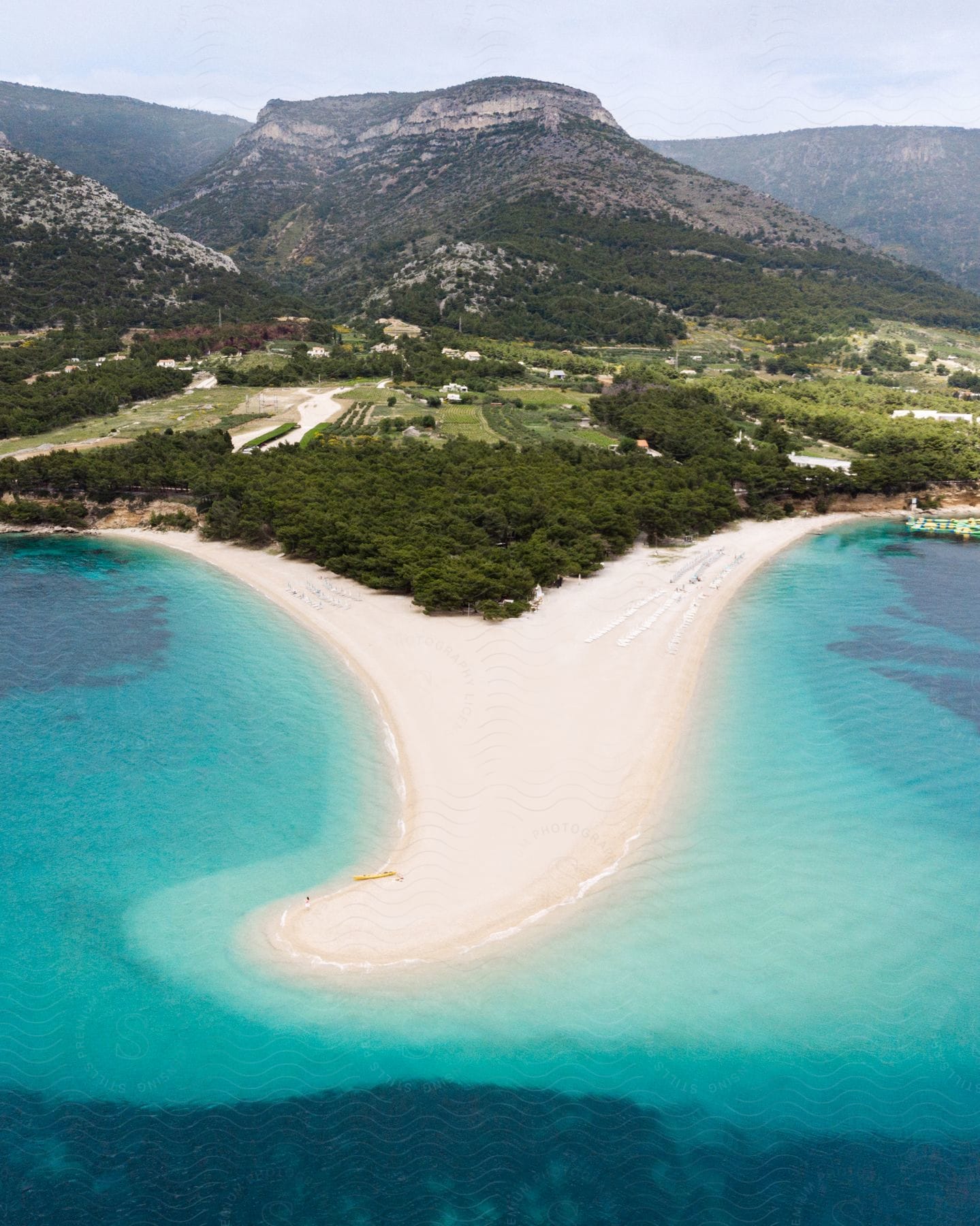 A beach stretches out into the clear blue water with trees and buildings inland and mountains in the distance