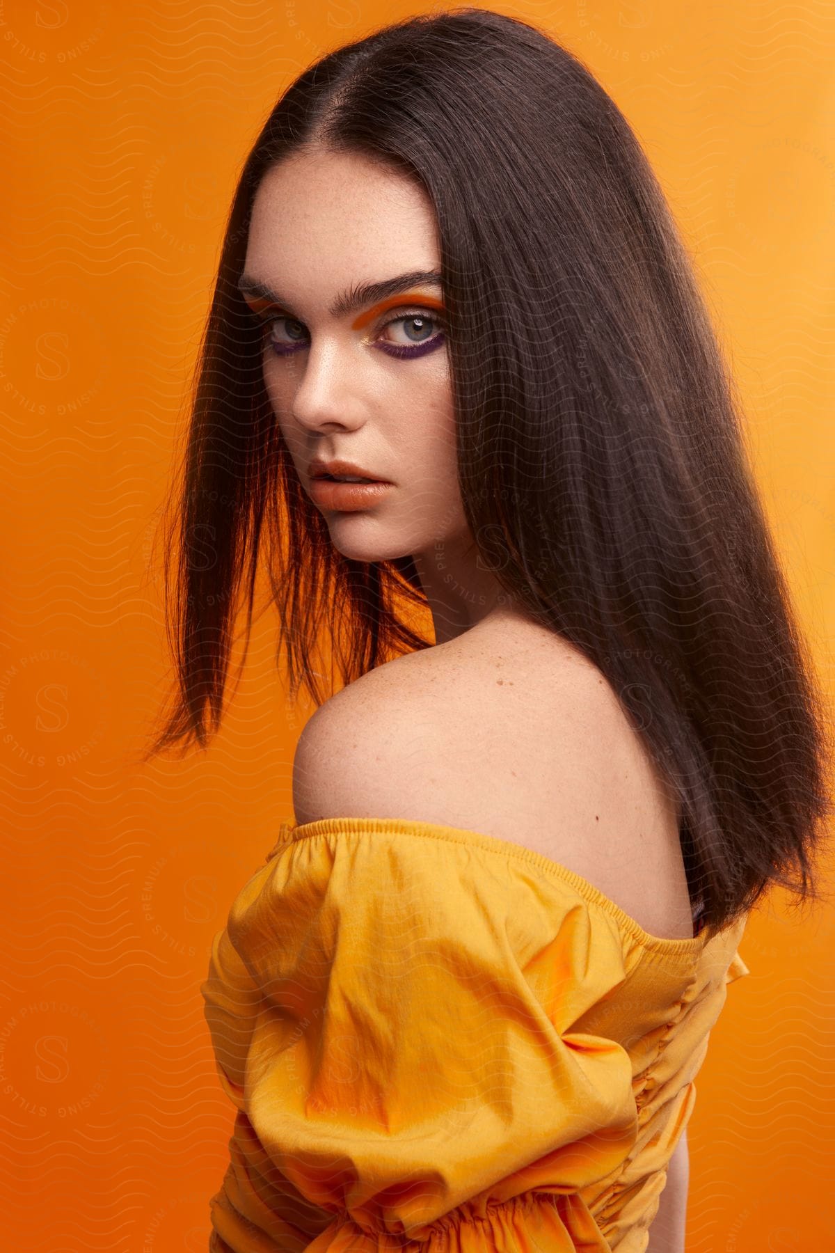 Green-eyed woman with brown shoulder-length hair and orange top with bare shoulder wears orange eyeshadow above and purple eyeshadow below, against an orange background.