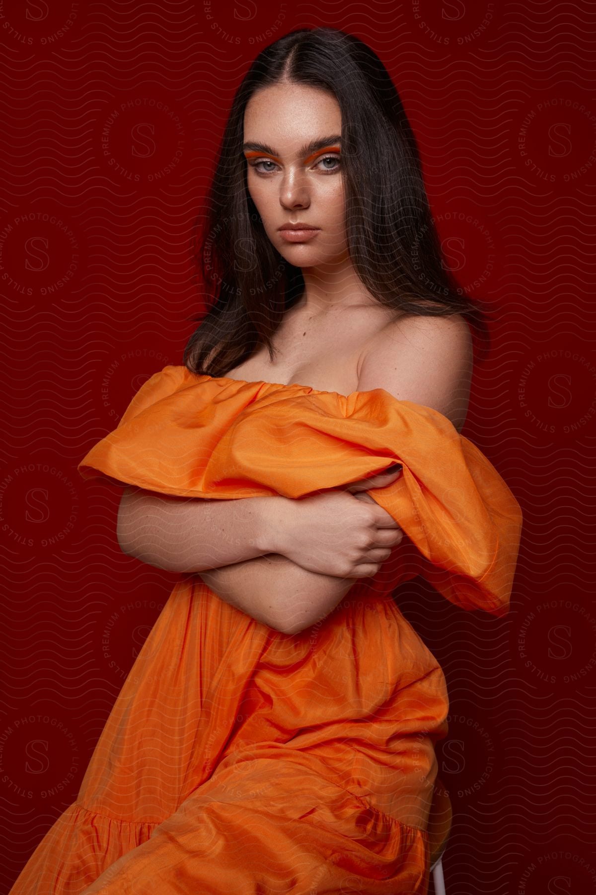 A woman wearing an orange dress is sitting on a chair in front of a burgundy red wall.