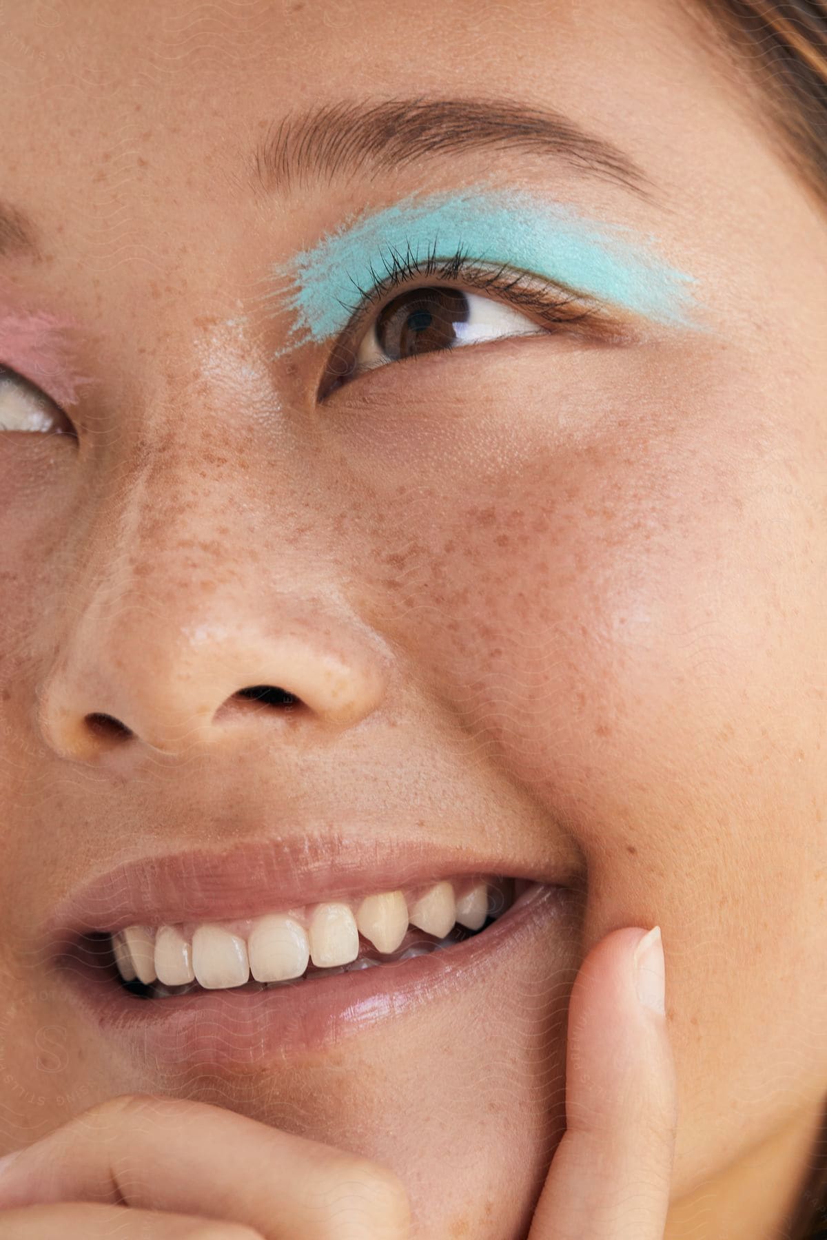 A girl wearing a stripe of light blue paint above her eye looks up as she smiles with her hand on her chin