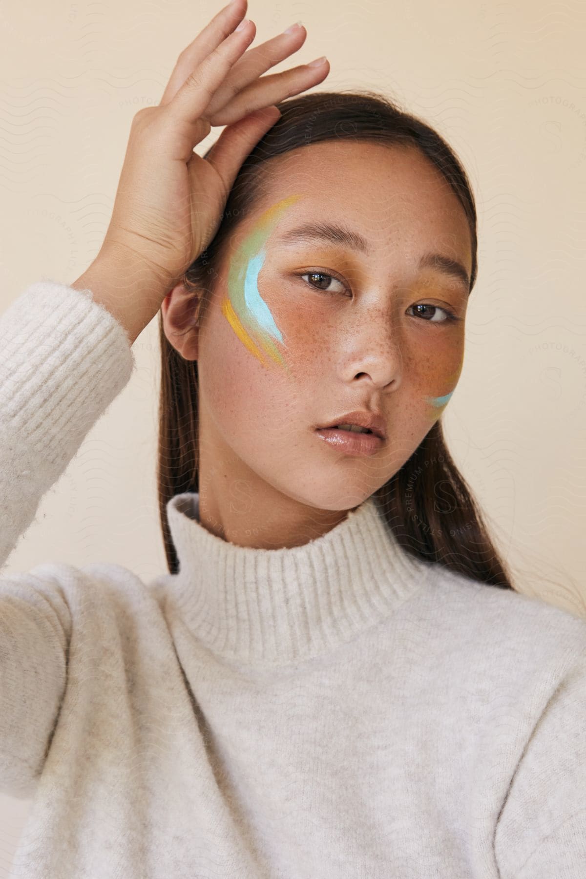 A woman with colored stripes on the side of her face holds her hand up as she looks ahead