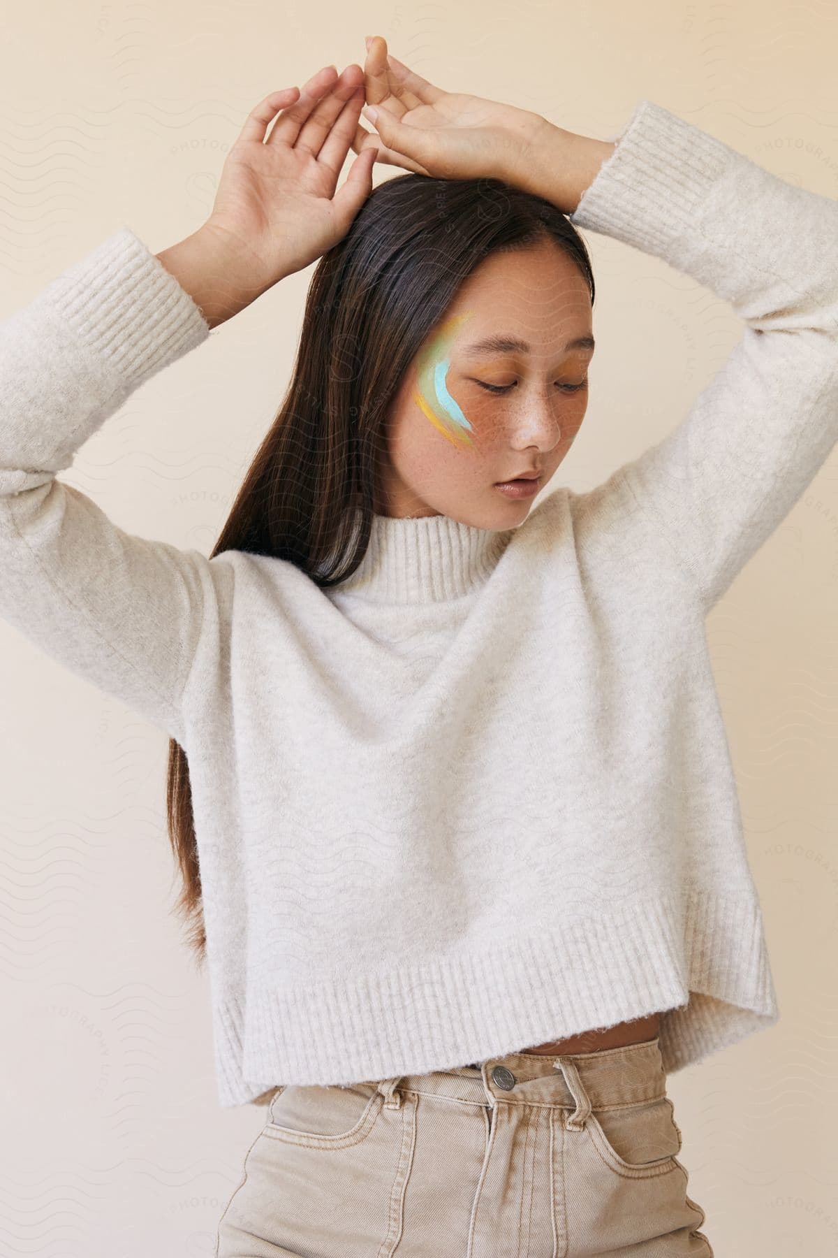 A Woman With Paint Stripes On The Side Of Her Face And Wearing A White Sweater With Her Arms Raised As She Looks Down