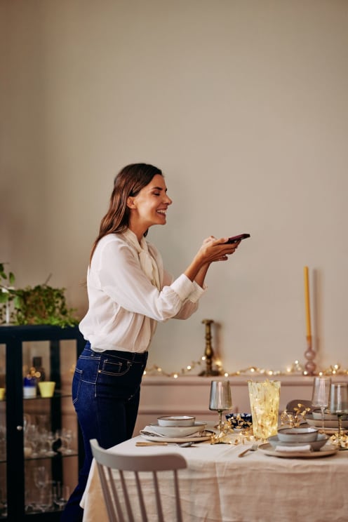 Woman With Brown Hair White Shirt And Blue Jeans Takes Photo Of Table With A Smartphone Laughing