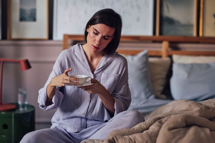 A Woman Is Sitting On Her Bed Holding A Cup Of Coffee