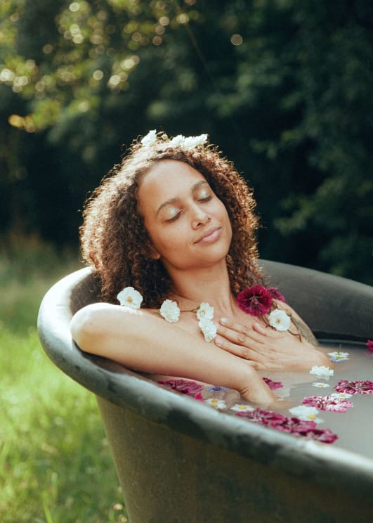 A Woman With Curly Hair Adorned In Flowers Relaxes Peacefully In An Outdoor Bathtub Filled With Flower Petals, Surrounded By Sunlight And Greenery