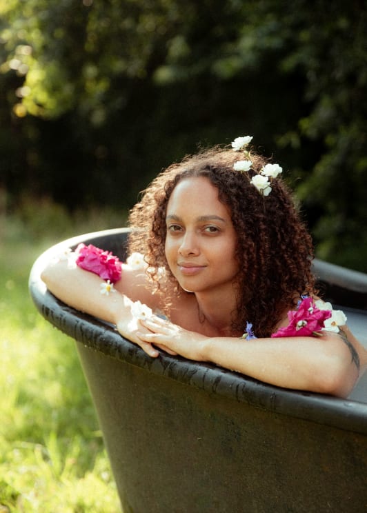 Woman Relaxing In An Outdoor Bathtub, With Flowers Adorning Her Hair And Arms, In A Natural And Serene Environment