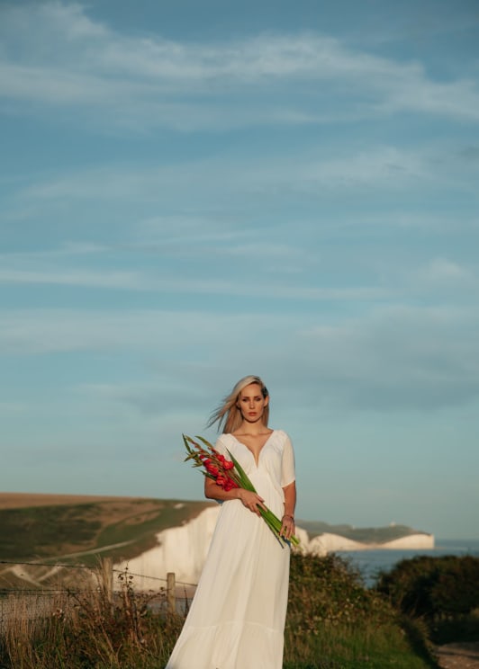 A Woman With Blonde Hair Wearing A Long White Dress Is Standing Along The Coast Holding A Bouquet Of Flowers