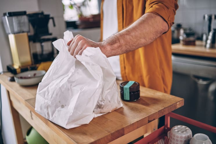 A Man Wearing A White Top And An Orange Jacket, Holding Two Bags Of Food On A Wooden Kitchen Counter