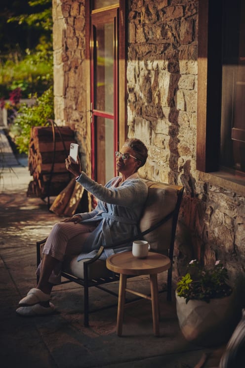 Black Woman Sits In Chair In Front Of Stone House Holding Iphone