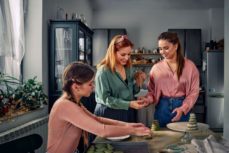A Mother With Red Hair, A Green Shirt, And A Blue Skirt, Along With Her Daughters Wearing Pink Shirts And Jeans, Make Cookies In The Kitchen, Sharing Laughter