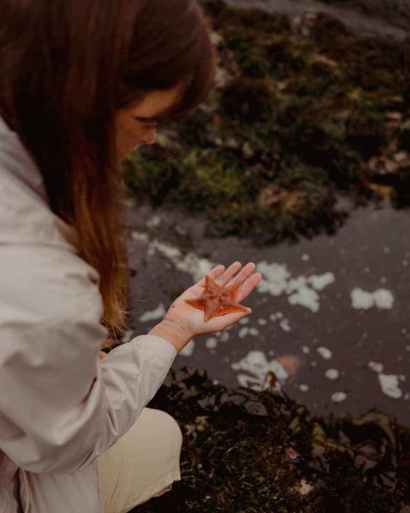 A Woman Crouching By The Water Holding A Starfish In The Palm Of Her Hand