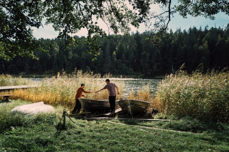 Two Male Friends Standing Together On The Riverbank Near Two Docked Row Boats