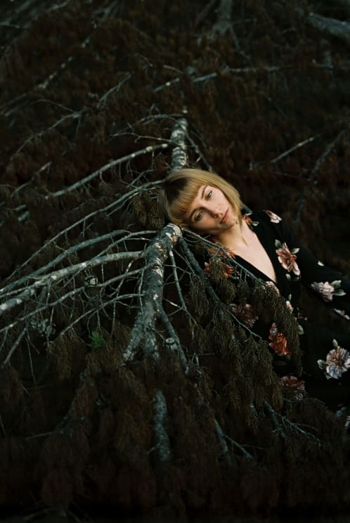 Blonde Model Wearing A Long Black Dress Leans Her Head Against A Fallen Tree Trunk