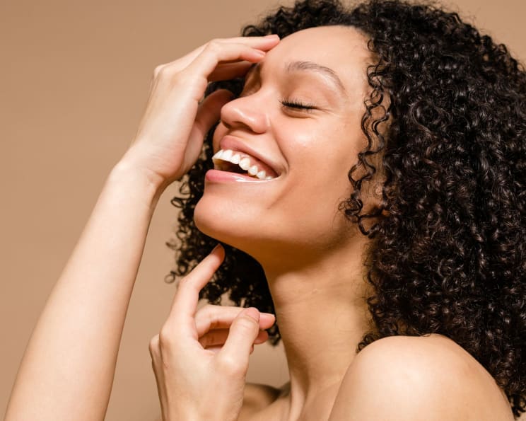 Side Portrait Of A Young Curly-haired Woman With Her Eyes Closed Giving A Beautiful Smile And With Her Hands On Her Face And Neck