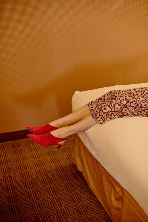 Woman Wearing Red Shoes And A Red Patterned Dress Lies On A Bed In A Room Decorated In Orange