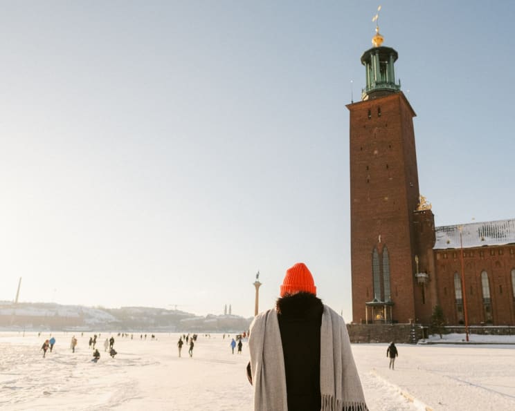 Woman Wearing A Red Hat A Gray Scarf And Black Coat Walks By Stockholm City Hall With People Scattered On The Snowy Ground
