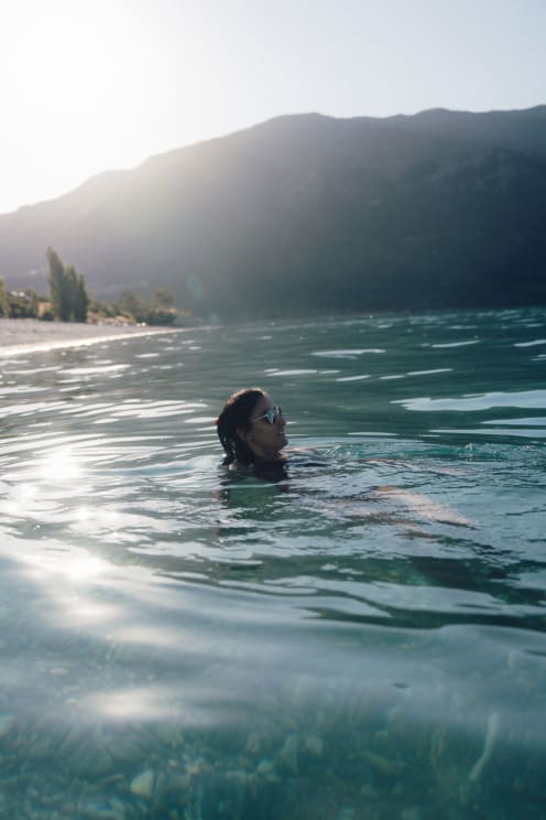 Woman Wearing Sunglasses In The Water Off The Coast With Mountains In The Distance