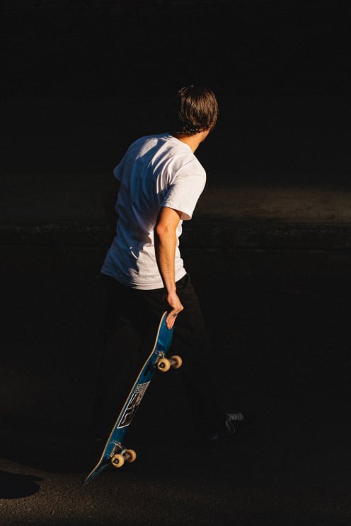 A Man Wearing A White T-shirt And Black Pants Walks Down The Street, Carrying A Blue Skateboard