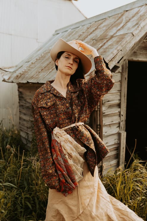 A Woman Holding A Hat, Wearing A Floral Shirt, Standing Near An Old Wooden Structure In Tall Grass