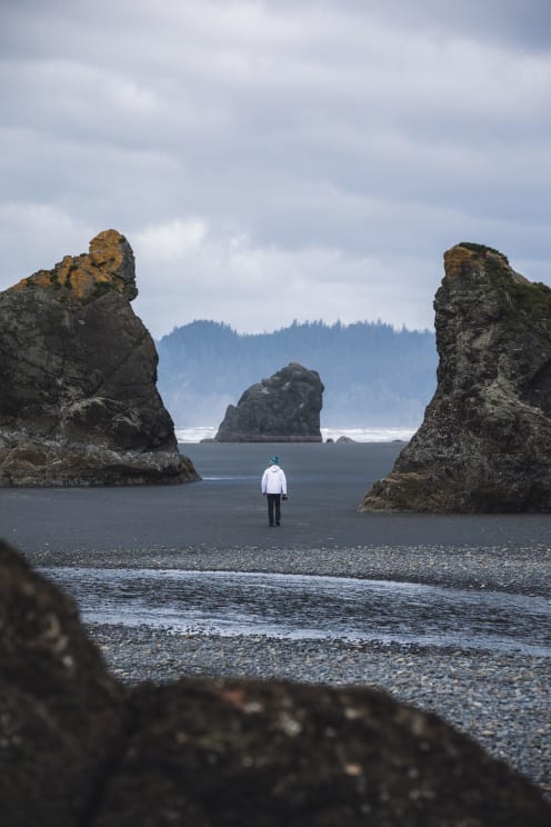 A Man, Sporting A Blue Hat, A White Coat, And Black Pants, Strolls Along Ruby Beach, Camera In Hand, Ready To Capture The Scenic Beauty Of The Coastline