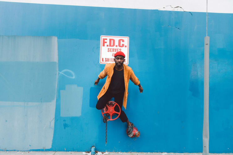 A Person Leaning Against A Blue Wall, Wearing Colorful Clothes, Including A Vibrant Yellow Jacket, A Black Blouse And Black Pants