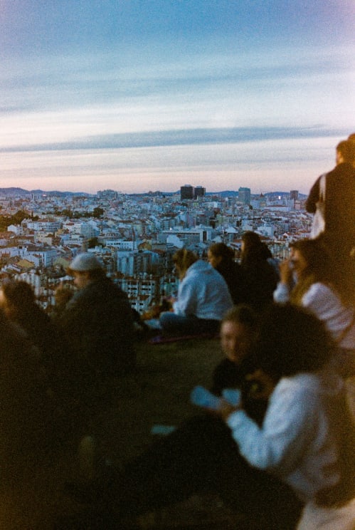 People Sit On The Ground Of A Hill Overlooking The City Against A Red Tinted Horizon