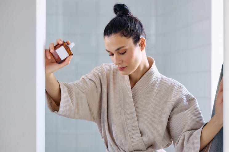 A Robed Woman Stands In A Bathroom While Holding A Bottle Of Skin Serum In Her Hand