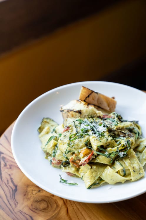 A White Plate Topped With Pesto Pasta And A Side Of Bread Sits On A Wooden Table