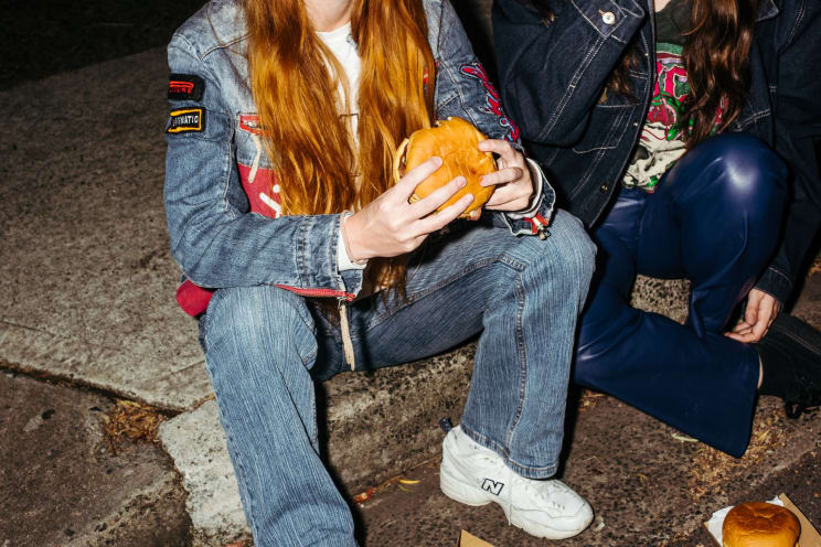 Woman With Red Hair, A Denim Jacket, Jeans, And White Sneakers Sits On The Curb Near A Woman Eating A Hamburger On The Street At Night