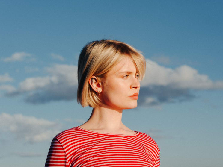 Woman With Short Blonde Hair Wearing A Red Striped Shirt Stands Outdoors Against A Blue Sky With White Clouds