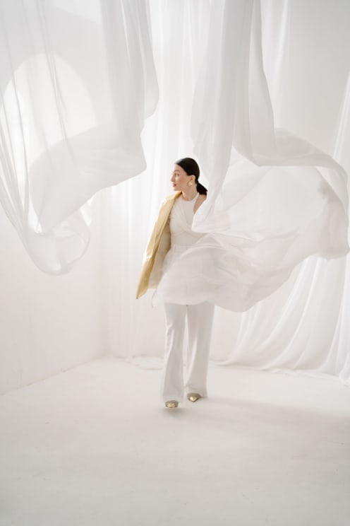 A Woman Wearing White Clothes And Gold Shoes, In A White Space With Flowing Curtains