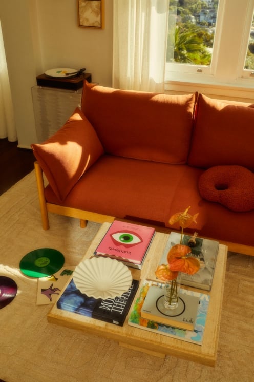 A Cozy Living Room With A Bright Orange Sofa, And Wooden Coffee Table With Books And A Plant On It