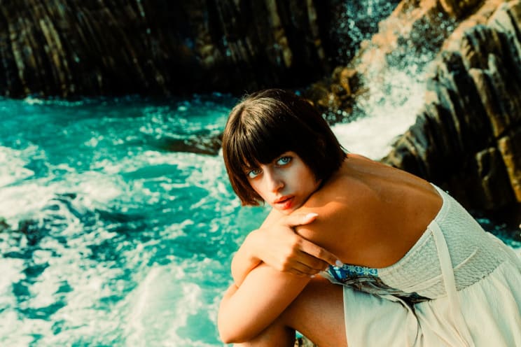 A Woman With Short Black Hair, Wearing A White Dress, Is Sitting Near A Body Of Water With Waves And Rocks In The Background