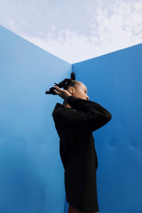 Model Woman Wearing A Long Black Coat With One Hand In Her Hair And Surrounded By Blue Walls