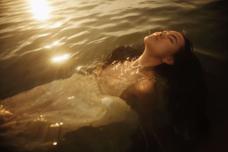 Young Woman Wearing A White Dress Is Lying In The Water Along The Coast With Her Eyes Closed As The Sun Shines Brightly Upon The Water