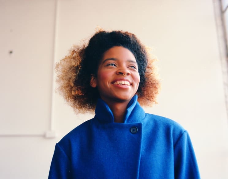 A Young Black Woman Smiles While Wearing A Blue Coat And A Gold Nose Ring