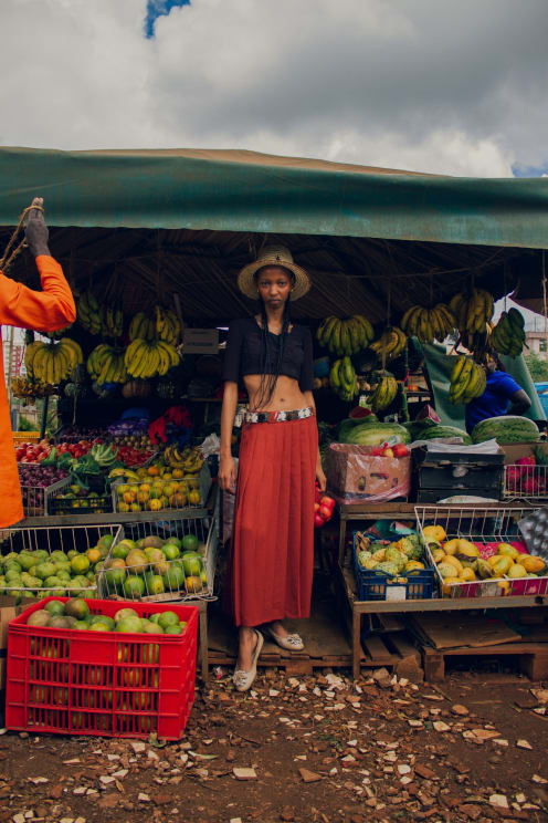 A Young Black Woman Stands In A Farmers Market While Wearing A Long, Red Skirt