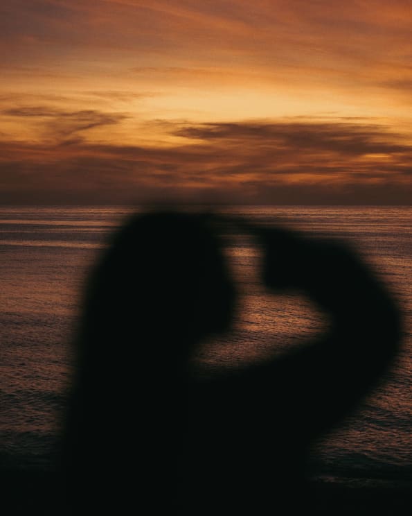 The Silhouette Of A Woman Holding Her Hand To Her Head While Overlooking The Ocean At Dusk