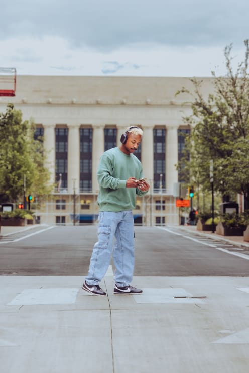 Man Wearing Headphones Stands On A City Street As He Looks At His Phone