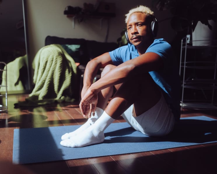 A Black Man With Blond Hair Sits On A Mat On A Living Room Floor While Wearing Headphones