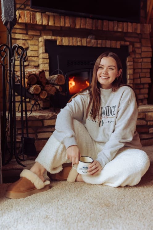 Woman In Pajamas Holding A Cup Sitting Next To A Fireplace And On A White Rug