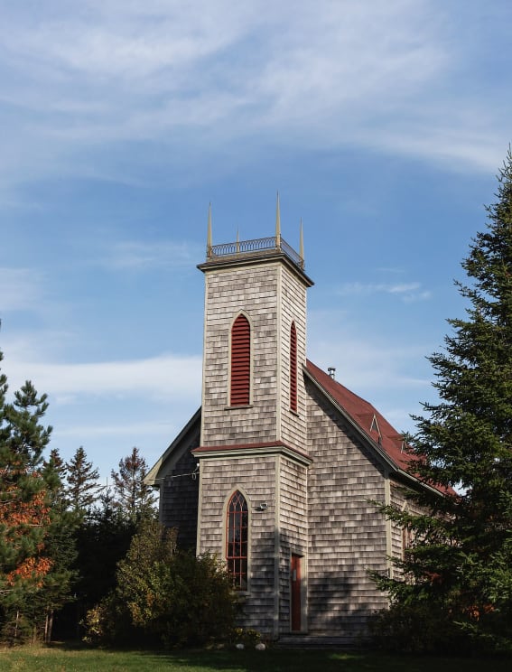 A Wood Shingle Church With Red Trim Sits In A Rural Area